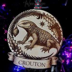May include: Wooden Christmas ornament with a lizard illustration. The round ornament features a detailed engraving of a lizard, holly, and a banner that reads "CROUTON". The ornament is suspended by a green and red ribbon.
