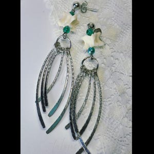 May include: A pair of silver-toned dangle earrings. Each earring features a small, white, bone-shaped charm, green beads, and multiple curved, textured metal strands. The earrings have a post-and-stud closure and are displayed on a white, lace-like surface.