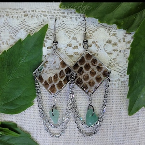 May include: A pair of dangle earrings with a diamond-shaped design. The earrings feature a snakeskin pattern encased in clear resin, with silver-colored chains and small, light green gemstone accents. The earrings hang from silver-colored ear wires.