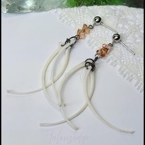 May include: Unique dangle earrings featuring bone-shaped elements. The earrings have a silver-tone post and a cluster of orange-colored crystals. The bone-shaped elements are white and curved, creating a distinctive and artistic design. The earrings are set against a white background.