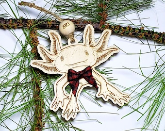 May include: A wooden Christmas ornament shaped like an axolotl wearing a red and black plaid bow tie. The ornament is hanging from a pine branch and has the year 2023 written on it.