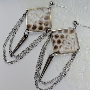 May include: A pair of silver-toned earrings with square, clear resin pendants containing a brown and white pattern. Each earring features a chain and a dark gray spike charm, creating a unique, edgy design. The earrings are displayed on a white, textured surface.
