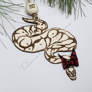 May include: A wooden Christmas ornament shaped like a snake with a red and black plaid bow. The ornament is decorated with a brown and white pattern and has the year 2022 written on it.