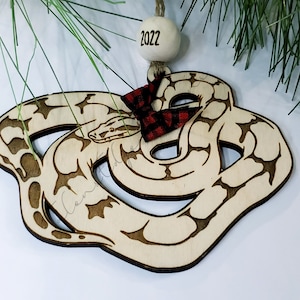 May include: A wooden Christmas ornament shaped like a snake with a red and black plaid bow. The ornament is decorated with a wood-burned design and has the year 2022 written on it.