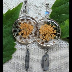 May include: A pair of handmade earrings featuring clear resin circles with dried yellow flowers and a textured background. Each earring has a silver wire and a gray crystal pendant. The earrings are displayed on a white lace surface.