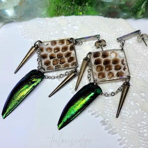 May include: Unique handmade earrings featuring square resin pendants with a reptile skin pattern, accented with gold-toned spikes and iridescent green beetle wings. The earrings are connected by silver chains and posts.