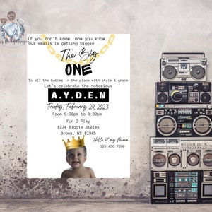 May include: A white invitation with gold accents for a first birthday party. The invitation features a gold chain with a crown and the text "The Big One" and "A.Y.D.E.N". The invitation also includes the date "Friday, February 24, 2023" and the time "From 5:30pm to 8:30pm". The invitation is decorated with a stack of vintage boomboxes.