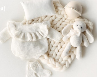 Puede incluir: Un conjunto de bebé color crema etiquetado como Set #1, que incluye un mameluco de manga larga con volantes, una almohada rectangular pequeña, un conejo de punto, un par de calcetines y una diadema sobre una manta de punto grueso.