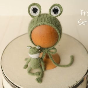 May include: A sage green knitted bonnet featuring two frog eyes with white and black centers, accompanied by a small matching knitted frog toy. The text "Frog Set #1" is visible in the upper right corner of the image.