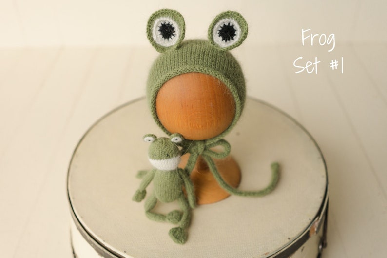 May include: A sage green knitted bonnet featuring two frog eyes with white and black centers, accompanied by a small matching knitted frog toy. The text "Frog Set #1" is visible in the upper right corner of the image.