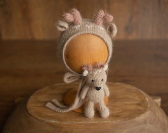 Puede incluir: Un gorro de punto marrón en forma de ciervo con orejas rosas y un juguete de punto marrón a juego en forma de ciervo. El gorro está sentado en un soporte de madera.
