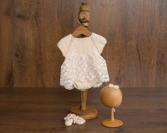 May include: A cream-colored knitted baby dress with a lace overlay, displayed on a wooden hanger with a wooden stand. Two small cream-colored booties are on the floor next to the stand. A wooden doll head with a white bow is on a separate wooden stand.