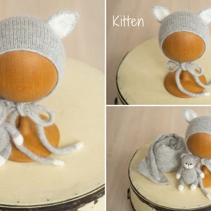 May include: A light gray knitted bonnet featuring pointed ears with white inner details, accompanied by a matching small gray and white knitted kitten toy and a long gray knitted wrap. The word Kitten is printed in white text in the corner.
