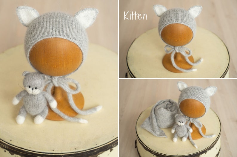 May include: A light gray knitted bonnet featuring pointed ears with white inner details, accompanied by a matching small gray and white knitted kitten toy and a long gray knitted wrap. The word Kitten is printed in white text in the corner.