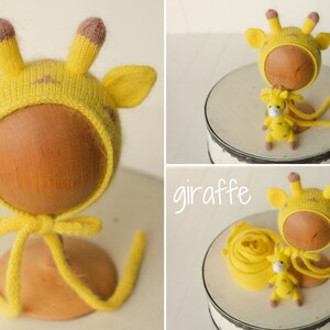 May include: A yellow knit bonnet featuring giraffe ears and brown-tipped horns, accompanied by a small matching giraffe plush toy. The set includes a yellow fabric wrap, and the word giraffe is written in white script text on the image.