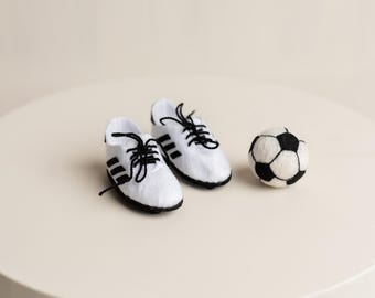 Puede incluir: Botas de fútbol blancas con rayas y cordones negros, junto a un balón de fútbol blanco y negro. Los artículos se muestran sobre una superficie neutra, evocando un tema deportivo.