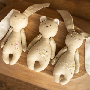 May include: Three handmade plush toys: a brown bear and two beige bunnies with embroidered flowers and a bee. The toys are lying on a wooden surface.