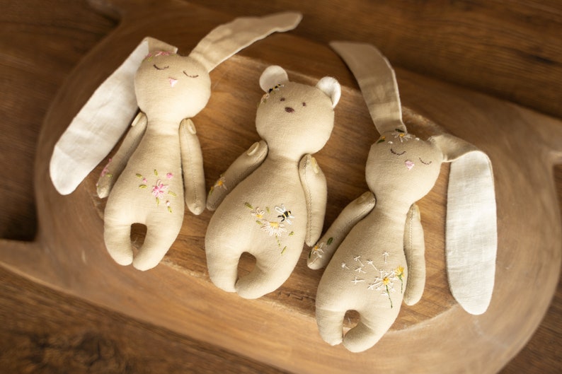 May include: Three handmade plush toys: a brown bear and two beige bunnies with embroidered flowers and a bee. The toys are lying on a wooden surface.