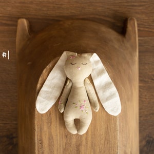 May include: A handmade beige bunny toy with white fabric ears and pink floral embroidery. The bunny is lying on a wooden surface.