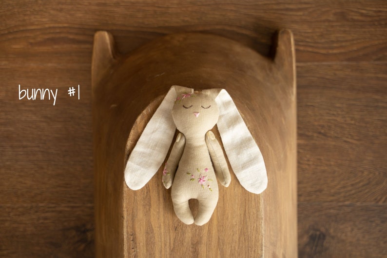 May include: A handmade beige bunny toy with white fabric ears and pink floral embroidery. The bunny is lying on a wooden surface.
