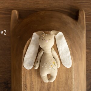 May include: A beige fabric bunny with embroidered flowers and closed eyes. The bunny is lying on a wooden surface.