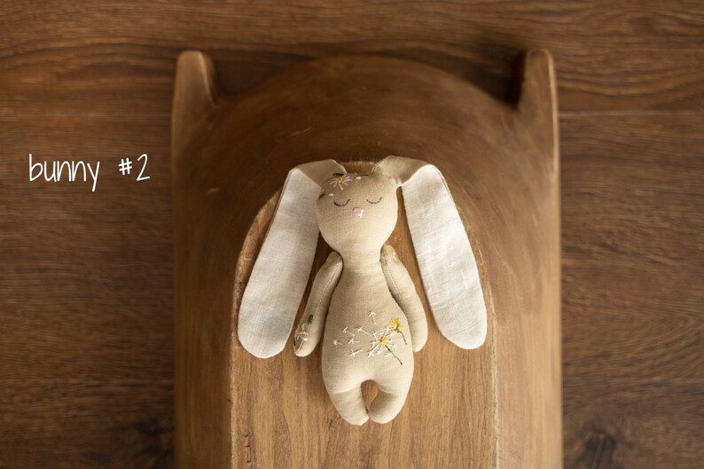 May include: A beige fabric bunny with embroidered flowers and closed eyes. The bunny is lying on a wooden surface.