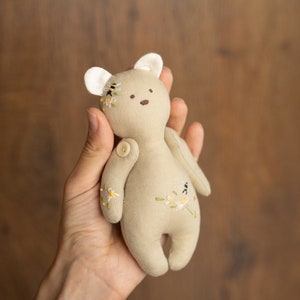 May include: A small, beige, handmade plush teddy bear with embroidered flowers and bees. The bear has a button nose and is held in a hand.