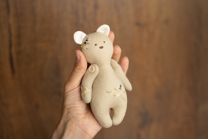 May include: A small, beige, handmade plush teddy bear with embroidered flowers and bees. The bear has a button nose and is held in a hand.