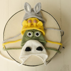 May include: A stack of four knitted animal-themed hats. The top hat is grey with rabbit ears, the second is yellow with giraffe horns, the third is green with large frog eyes, and the bottom hat is white with two black patches resembling a cow nose.