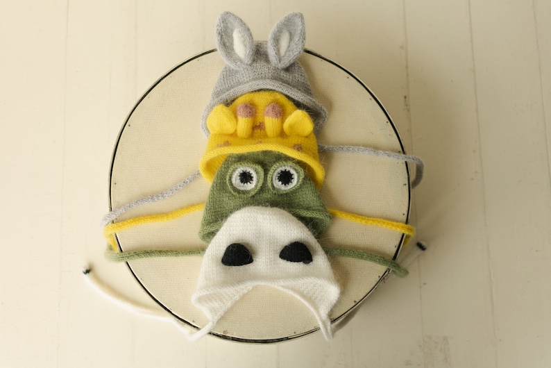 May include: A stack of four knitted animal-themed hats. The top hat is grey with rabbit ears, the second is yellow with giraffe horns, the third is green with large frog eyes, and the bottom hat is white with two black patches resembling a cow nose.