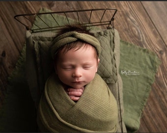 Puede incluir: Un bebé recién nacido duerme envuelto en una manta verde y una diadema verde en una pequeña cama de metal. El bebé está acostado sobre una almohada y una manta verdes. El fondo es un suelo de madera.