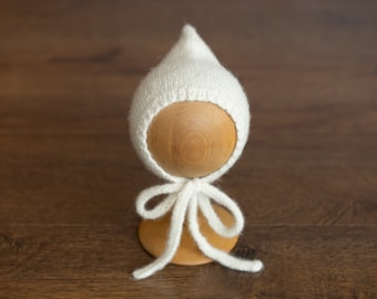 Puede incluir: Un gorro de bebé de punto blanco con la parte superior puntiaguda y una larga corbata. El gorro se muestra en una cabeza de maniquí de madera.