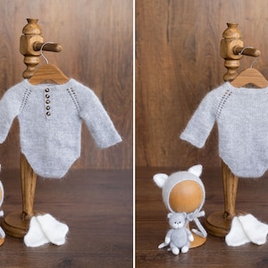 May include: A light gray long-sleeve knit bodysuit with a button-front placket and raglan sleeves. The set includes a matching gray knit bonnet with cat ears, a small gray stuffed cat toy, and a pair of white knit booties on a wooden stand.