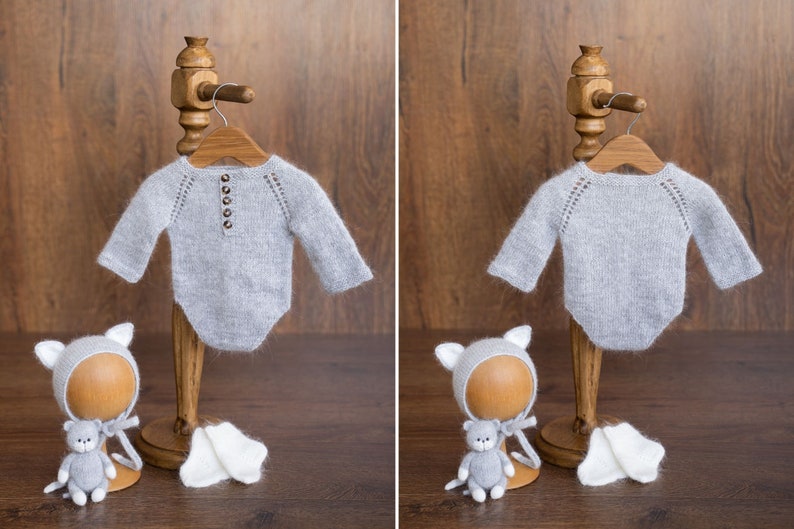May include: A light gray long-sleeve knit bodysuit with a button-front placket and raglan sleeves. The set includes a matching gray knit bonnet with cat ears, a small gray stuffed cat toy, and a pair of white knit booties on a wooden stand.