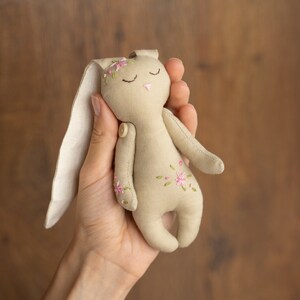May include: A beige fabric bunny with embroidered pink flowers and closed eyes. The bunny is held in a hand.