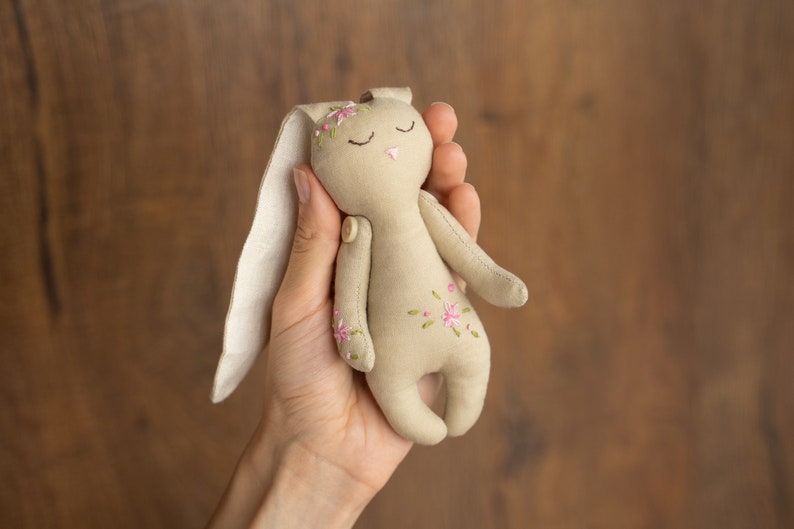 May include: A beige fabric bunny with embroidered pink flowers and closed eyes. The bunny is held in a hand.