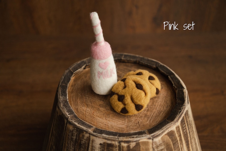 Puede incluir: Una botella de leche rosa y blanca afieltrada con una parte superior rosa y un coraz&oacute;n que dice "MILK" junto a dos galletas con chispas de chocolate. Los art&iacute;culos est&aacute;n sobre una superficie de madera con el texto "Pink set".