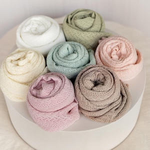 May include: Seven rolled baby blankets in various pastel colors, including white, cream, sage green, light blue, and pink. The blankets are arranged on a white circular platform, showcasing their delicate, open-weave texture.