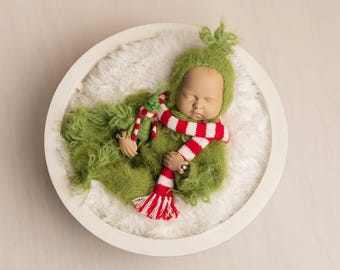 Puede incluir: Un bebé recién nacido con un disfraz verde con el tema del Grinch, que incluye un body y un gorro peludos, y una bufanda a rayas rojas y blancas. El bebé está acurrucado en una cama circular blanca y esponjosa.