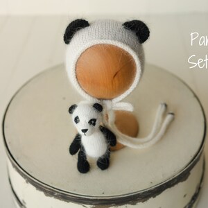 May include: A white and black knitted panda-themed bonnet with matching ears and a small knitted panda toy. The set is labeled Panda Set #1. The bonnet features long ties and the toy has black patches around the eyes, ears, arms, and legs.
