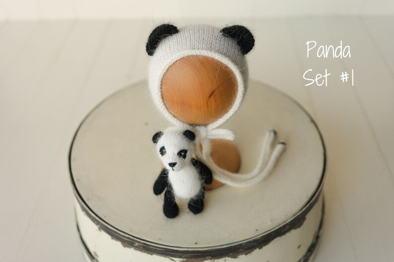 May include: A white and black knitted panda-themed bonnet with matching ears and a small knitted panda toy. The set is labeled Panda Set #1. The bonnet features long ties and the toy has black patches around the eyes, ears, arms, and legs.