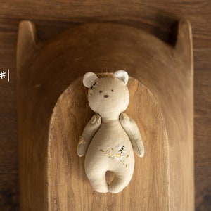 May include: A beige plush teddy bear with embroidered flowers and bees. The bear has white ears and is lying in a wooden bowl.