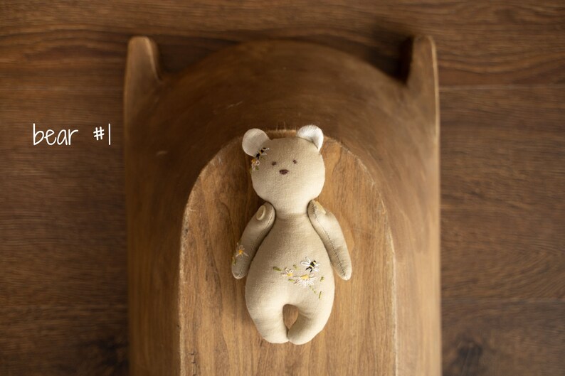 May include: A beige plush teddy bear with embroidered flowers and bees. The bear has white ears and is lying in a wooden bowl.