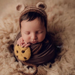 Puede incluir: Un beb&eacute; reci&eacute;n nacido envuelto en una envoltura marr&oacute;n, con un gorro de orejas de oso marr&oacute;n y beige. El beb&eacute; sostiene una galleta de fieltro con chispas de chocolate marr&oacute;n. El beb&eacute; est&aacute; acostado sobre una superficie esponjosa de color marr&oacute;n claro.