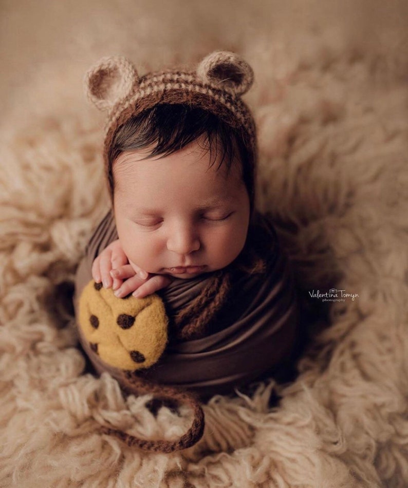 Puede incluir: Un beb&eacute; reci&eacute;n nacido envuelto en una envoltura marr&oacute;n, con un gorro de orejas de oso marr&oacute;n y beige. El beb&eacute; sostiene una galleta de fieltro con chispas de chocolate marr&oacute;n. El beb&eacute; est&aacute; acostado sobre una superficie esponjosa de color marr&oacute;n claro.