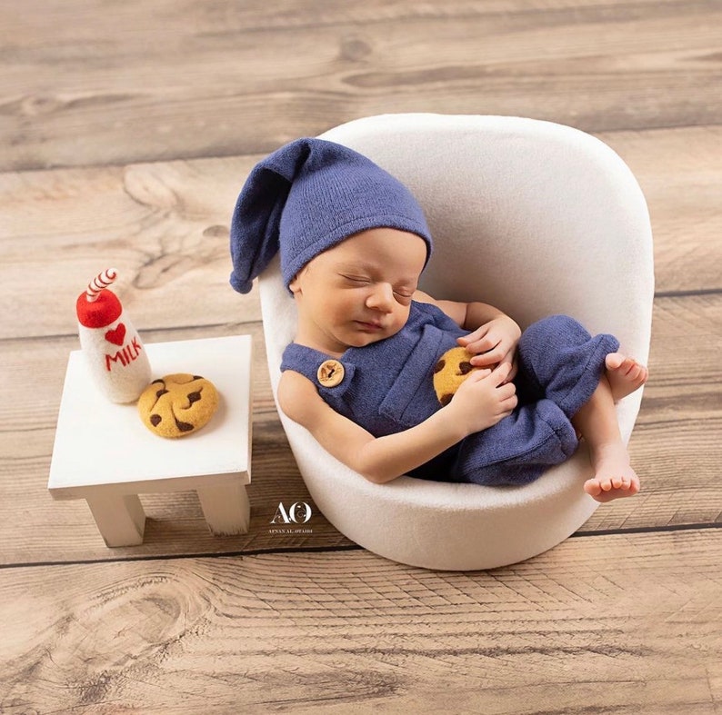 Puede incluir: Un beb&eacute; reci&eacute;n nacido con gorro y peto de punto azul, sosteniendo una galleta, descansando en una silla blanca. Una peque&ntilde;a mesa blanca sostiene una botella de leche de fieltro y una galleta. La escena est&aacute; ambientada en un suelo de madera.