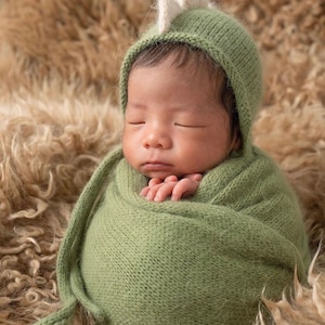 May include: A newborn infant wrapped in a sage green knitted swaddle blanket, wearing a matching sage green knitted bonnet that features a small, upright, cream-colored dinosaur-style crest on the top of the head, with the eyes closed.