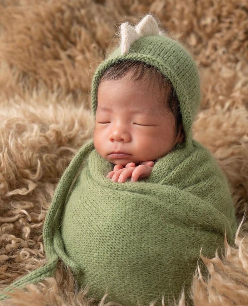 May include: A newborn infant wrapped in a sage green knitted swaddle blanket, wearing a matching sage green knitted bonnet that features a small, upright, cream-colored dinosaur-style crest on the top of the head, with the eyes closed.