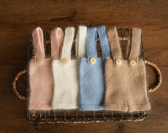 Puede incluir: Un conjunto de cinco petos de bebé tejidos en una cesta de alambre. Los petos son de color rosa, blanco, azul y marrón, cada uno con cierres de botón de madera. La textura suave y esponjosa sugiere un artículo acogedor y hecho a mano.