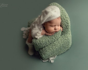 Puede incluir: Un bebé recién nacido que lleva un gorro blanco de punto con detalles de perlas está durmiendo en una silla verde peluda. El bebé lleva un atuendo de punto blanco y tiene una expresión suave y pacífica.